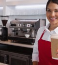 Happy young barista offering cup of coffee to go smiling at came