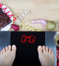 Digital scales with woman feet on them and sign"OMG!" surrounded by christmas decorations sweets and alcohol. Demonstrates consequences of surfeit and eating unhealthy food during Christmas holidays.